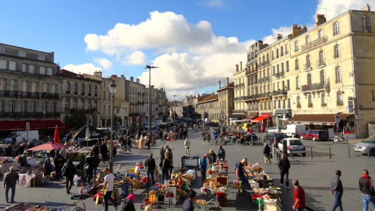 De nombreux étals de brocante installés sur une place urbaine ensoleillée entourée de bâtiments anciens.