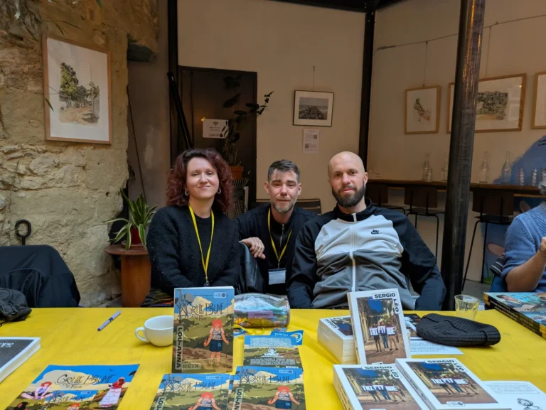 Trois personnes assises derrière une table jaune présentant des livres dans un espace d'art.