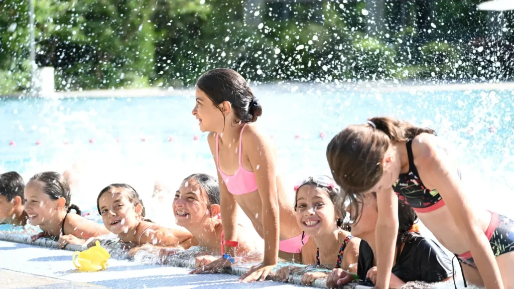 Des enfants sourient au bord d'une piscine ensoleillée, entourés par de nombreuses éclaboussures d'eau.