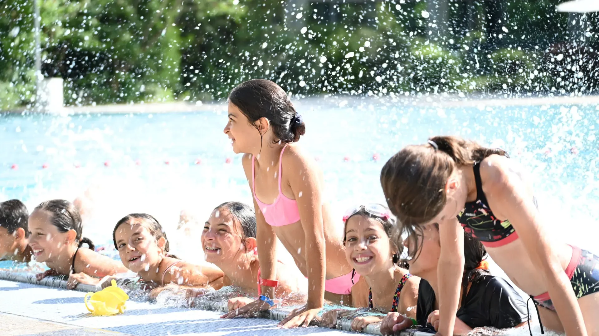 Des enfants sourient au bord d'une piscine ensoleillée, entourés par de nombreuses éclaboussures d'eau.
