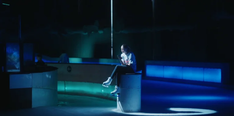 Patineuse assise sur un pilier utilisant son téléphone dans une patinoire aux lumières bleues.