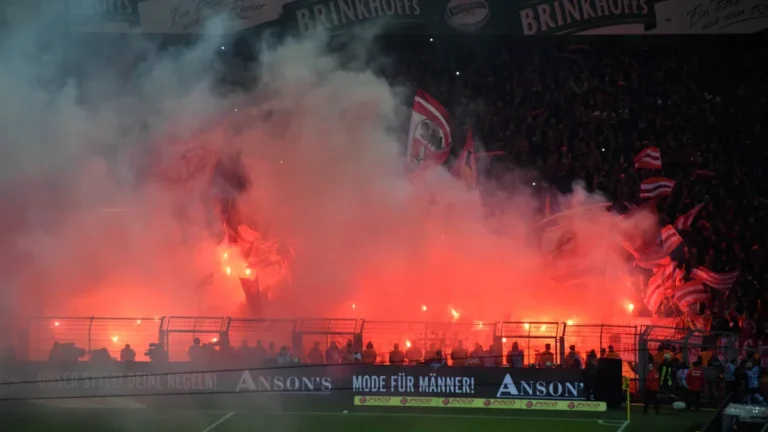 Supporters de football célébrant avec des fumigènes rouges dans un stade en fête.