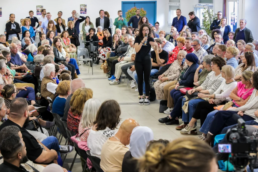 Jeune femme au micro s'adressant à une foule assise lors d'une assemblée citoyenne en intérieur.