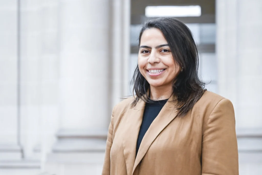 Portrait d'une femme souriante aux cheveux foncés portant un blazer marron devant des colonnes floues.