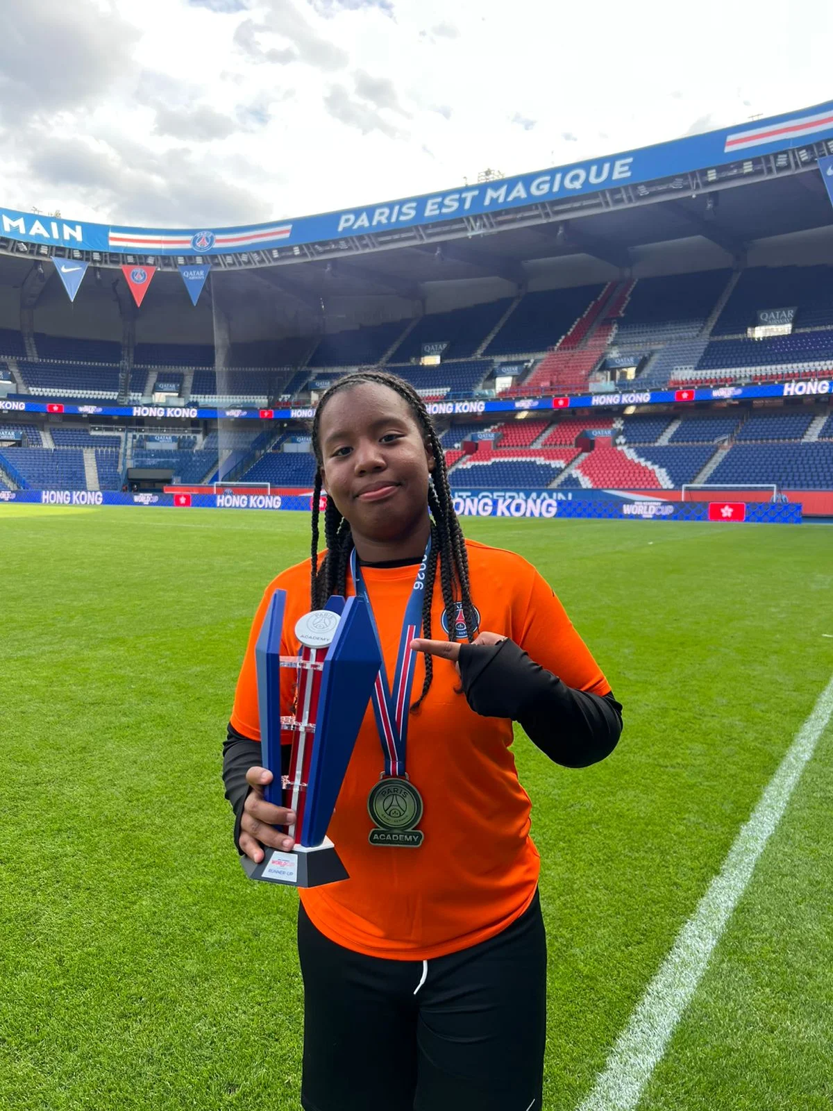 Jeune fille en maillot orange tenant un trophée et montrant sa médaille au milieu d'un stade.