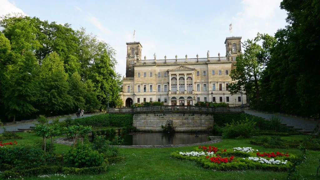 Un imposant bâtiment classique surplombant un parc urbain avec un bassin d'eau et des fleurs.