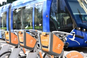 Plusieurs vélos de location Vélomagg orange garés devant un tramway bleu en circulation à Montpellier.
