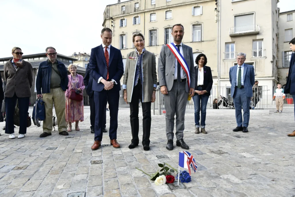 Officiels français et britanniques recueillis devant un dépôt de fleurs et de petits drapeaux au sol.