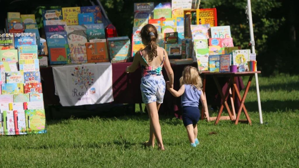 Deux jeunes enfants marchant vers un étalage de livres pour enfants dans un parc ensoleillé.