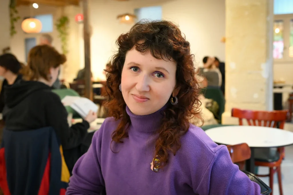 Une femme aux cheveux bouclés portant un col roulé violet sourit dans un café animé.