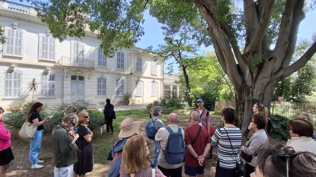 Un groupe de visiteurs écoute un guide devant une grande demeure bourgeoise aux volets bleus.