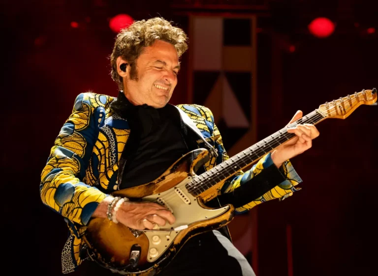 Un guitariste souriant portant une veste colorée joue d'une guitare électrique sur une scène.