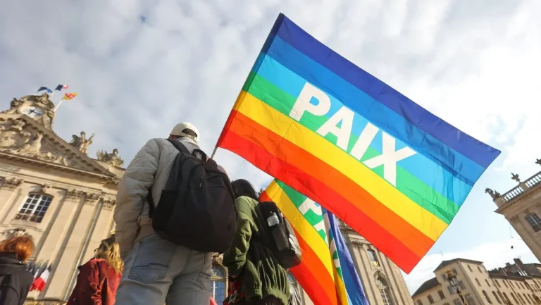 Un drapeau arc-en-ciel marqué « PAIX » tenu par des manifestants devant un bâtiment historique ensoleillé.