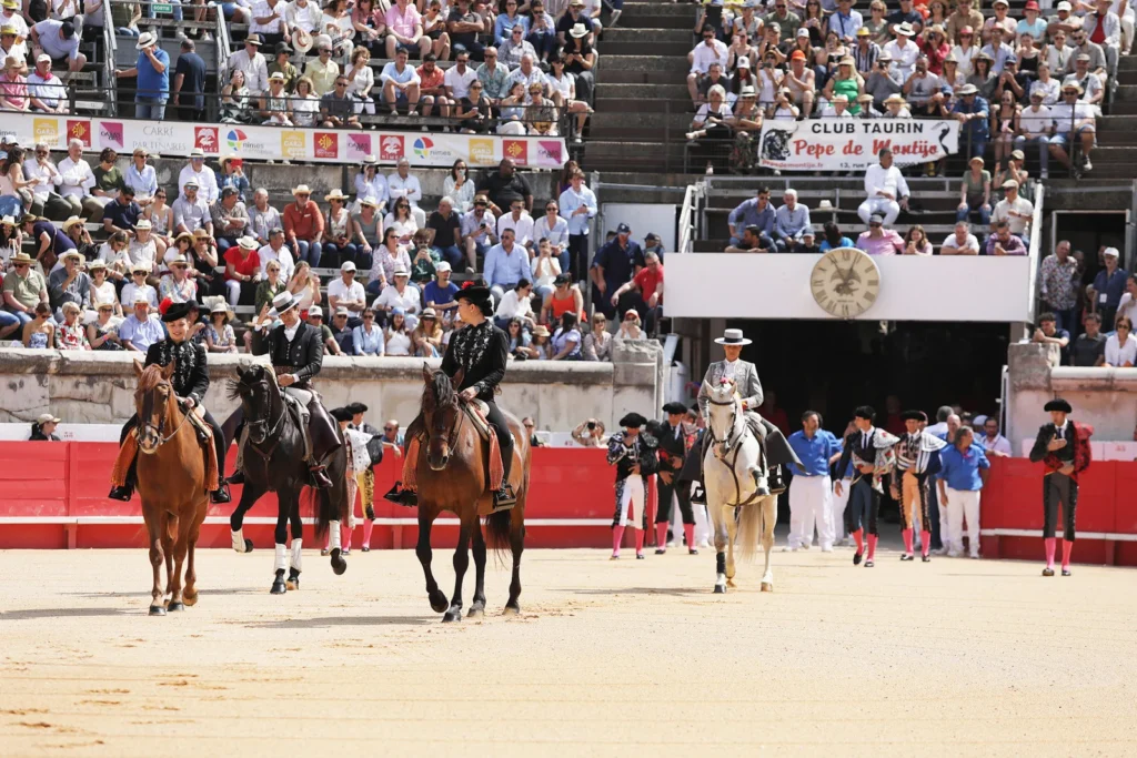 Quatre cavaliers en costume traditionnel défilent sur le sable d'une arène comble lors d'une feria.