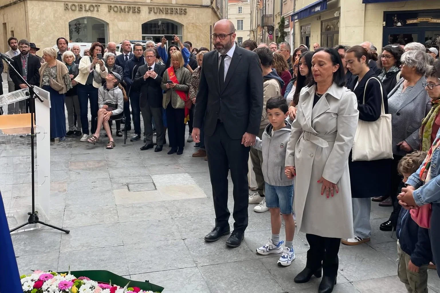Un homme, un jeune garçon et une femme se tiennent la main devant une foule recueillie.