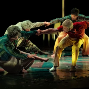 Quatre artistes en costumes colorés s'agrippent les mains lors d'une performance chorégraphique sur scène.