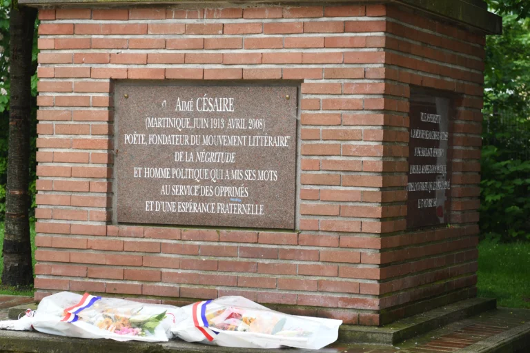 Plaque commémorative d'Aimé Césaire sur un mur de briques avec deux bouquets de fleurs déposés devant.