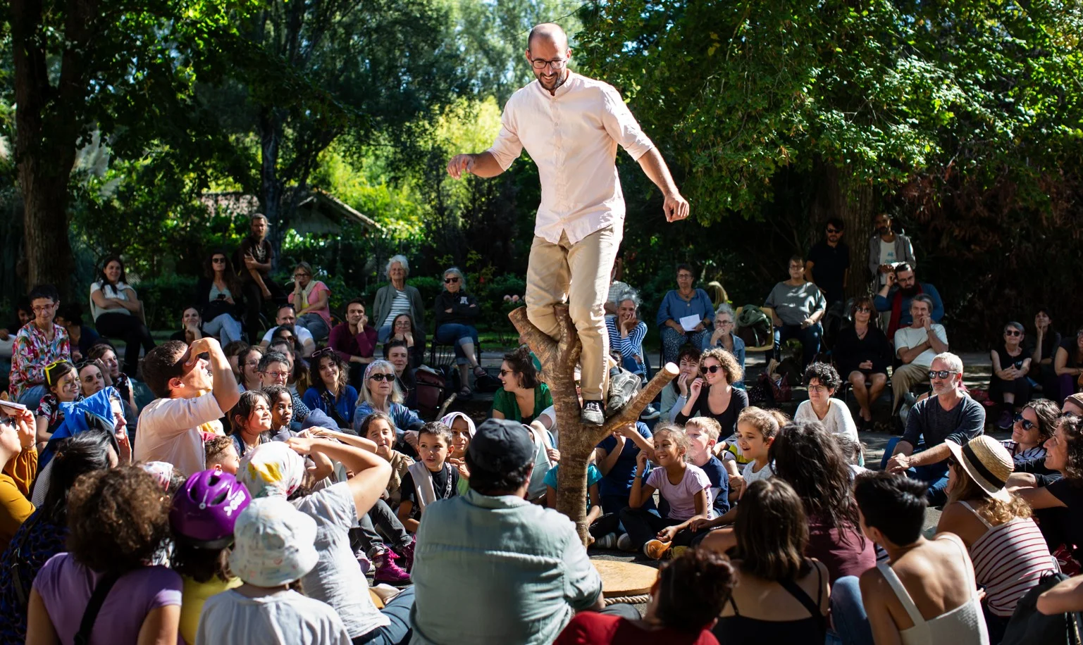 Un homme en équilibre sur une branche d'arbre sculptée devant un public assis en extérieur.