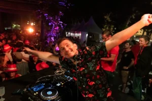 DJ souriant en chemise à fleurs bras écartés derrière ses platines lors d'un événement nocturne.