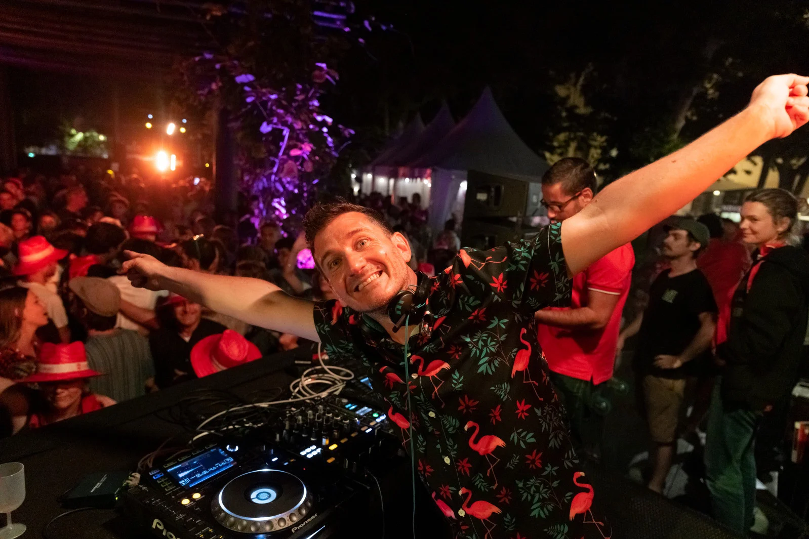 DJ souriant en chemise à fleurs bras écartés derrière ses platines lors d'un événement nocturne.