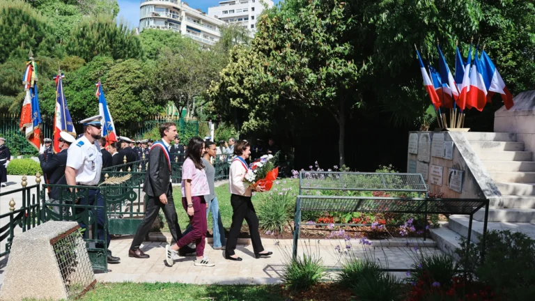 Un élu et une femme portant des fleurs marchent vers un monument pavoisé de drapeaux français.