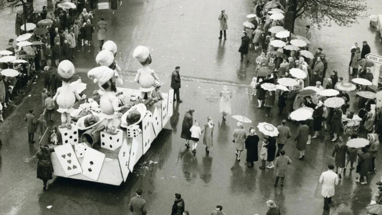Char de parade avec cartes et dés géants dans une rue pluvieuse remplie de parapluies.