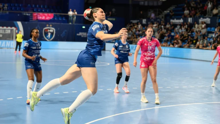 Une handballeuse en tenue bleue effectue un saut spectaculaire pour tirer vers le but.