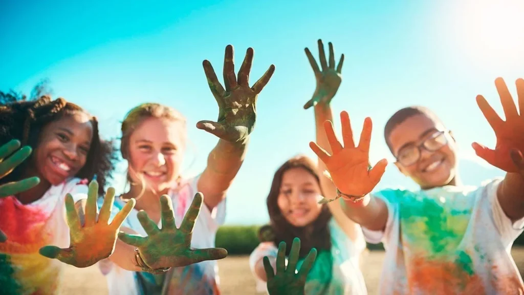 Quatre enfants souriants montrent leurs mains couvertes de peinture colorée sous un ciel ensoleillé.
