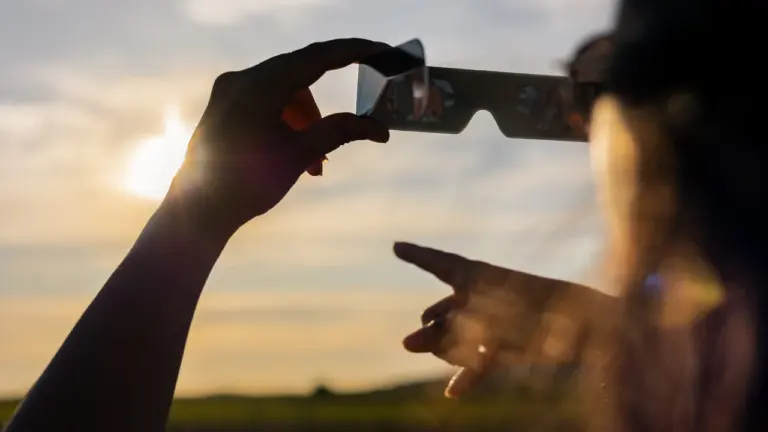 Une personne tient des lunettes d'éclipse solaire devant le soleil couchant à l'horizon.