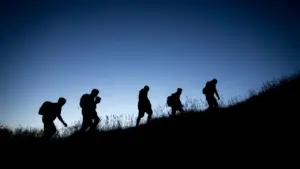 Silhouettes de randonneurs marchant sur une colline herbeuse sous un ciel bleu dégagé.
